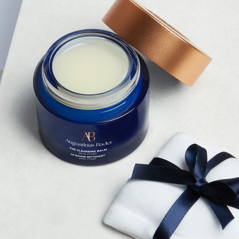 Product shot of 'The Cleansing Balm' by Augustinus Bader, which is displayed with its lid off on a table, next to a flannel which is wrapped in a bow