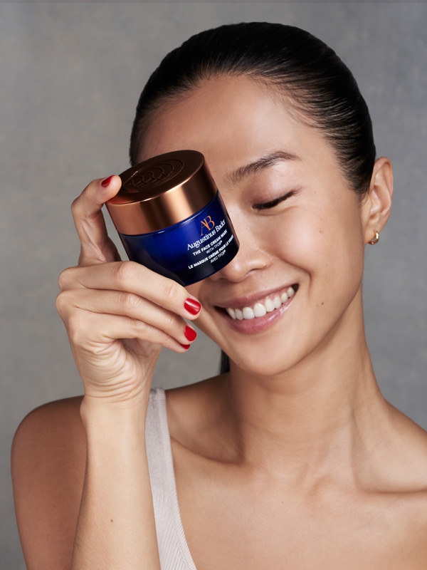 A model holds the Face Cream Mask by Augustinus Bader up to her face while smiling broadly