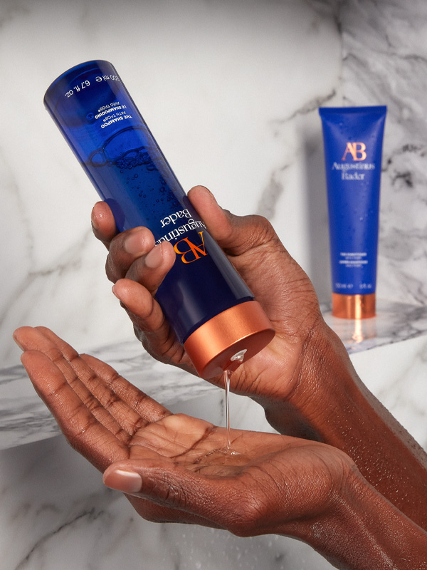 A model squeezes 'The Shampoo' by Augustinus Bader into her hands in the shower, with 'The Conditioner' displayed in the background