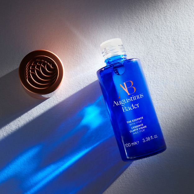 Product shot of 'The Essence' against a textured white background. Light shines through the transparent product and casts blue light from the bottle across the background